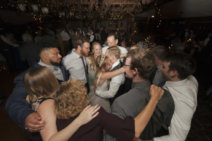 wedding event A couple in wedding attire embrace. Surrounding them in a tight circle are guests with their arms around each other.