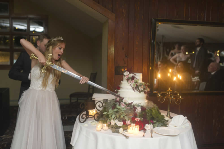wedding event A bride stands ready to battle in front of a table with a wedding cake and decorated in flowers and candles. She holds a long sword and is preparing to cut the cake.