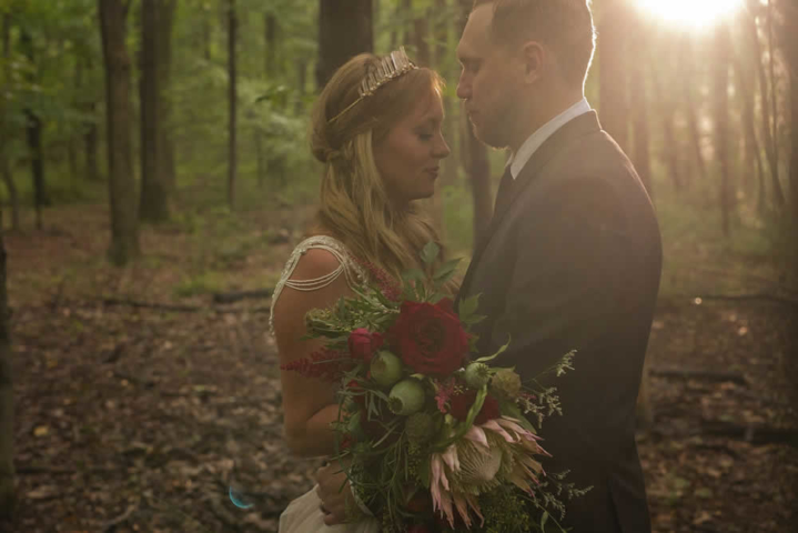 wedding event A couple in wedding attire at dusk in the forest. They are facing each other with eyes closed. A large bouquet of red and pink flowers are held in front of them.