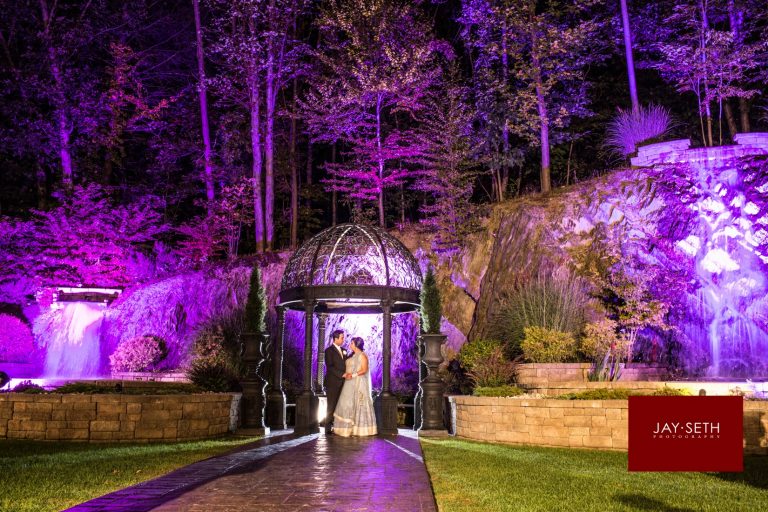 A couple in wedding attire stare at each other in front of an outdoor altar. It is nighttime and they are alone. The rocks and trees behind them are lit in purple lights.