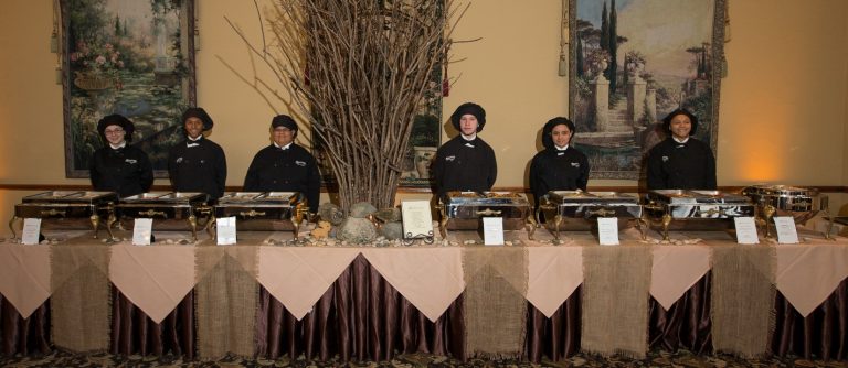 A long table dressed in maroon and tan linens with multiple serving trays of buffet style food. Six servers stand behind the table with black chef uniform and hats.