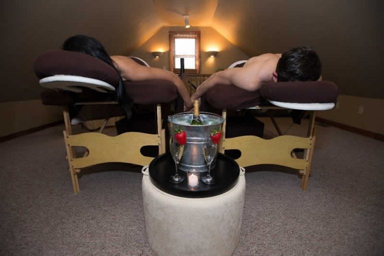 A couple are face down on massage beds while holding hands. A bucket of champagne is in between them.