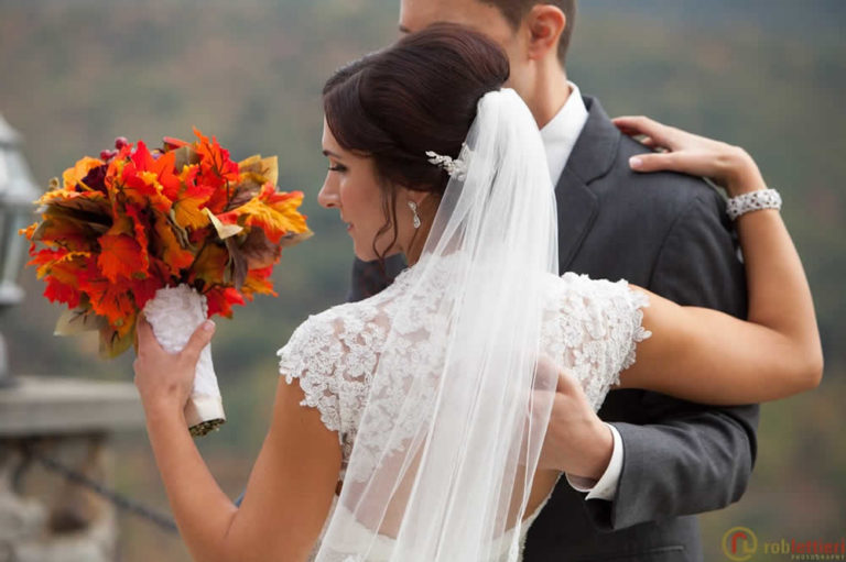 A bride holds a bouquet of autumn colored leaves. Her arms is around her groom.