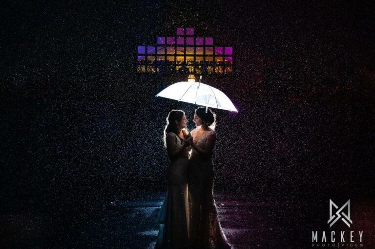 Two brides stand in the rain beneath an umbrella